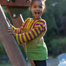 Little girl on a playground wearing sweater in colorful stripes. Yoke has square buttoned placket. Sleeves are long and full.