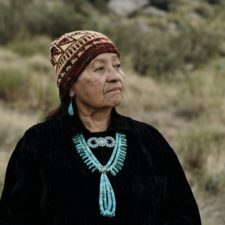 Older woman wearing elaborate turquoise jewelry models beanie with geometric colorwork.
