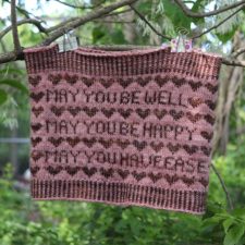 Two-color cowl is on a clothesline. The cowl has a hearts motif and reads, May you be happy. May you be well. May you have ease.