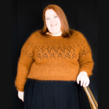 Victoria is wearing a pullover with a simple lace yoke with a chevron pattern. The yarn has a slight halo, and the tawny color matches her hair. Background and the rest of her outfit are black.