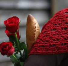 Woman wears lacy shawl with back to camera. In her arms are roses and French bread.