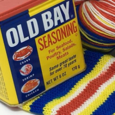 A can of Old Bay Seasoning in primary colors and matching striped sock yarn knitted up.