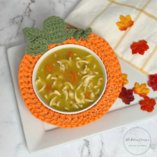Bowl of noodle soup rests inside crocheted bowl cozy in the form of a pumpkin.