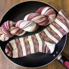 Skein and striped sock in a fry pan. Stripes look like bacon. Off-white stripes contain yellow egg yolk colored dots.