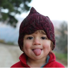 Kid sticking his tongue out, wearing hat with wavy hem and two points on top.