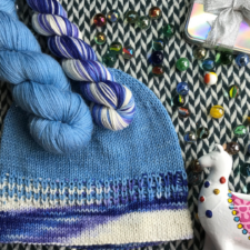 Two skeins of yarn, a llama figurine, some glass marbles and a mainly stockinette beanie made with a solid and a variegated yarn. The variegated yarn is at the brim and pools beautifully.