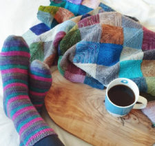 Mitered square blanket, and cup of coffee are on a table. Feet wearing knitted socks are resting on the table.