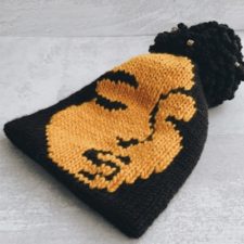 Two-color beanie with portrait of woman’s face. Top has a large pom-pom.