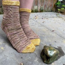 Socks with contrasting toes and cuff tops have a cable down the side. Model is standing next to what seems to be a brass frog.