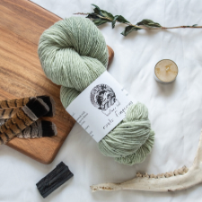 Pale green woolen-spun fingering yarn arranged with dried plants, a jawbone, a candle, feathers and charred wood.