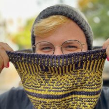 Smiling designer holds up colorwork cowl with intricate geometric colorwork.