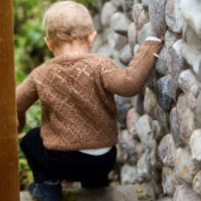 Small child walks down wooden stairs next to cobbled rock wall. Cardigan is longsleeve with a lattice and lace back, shown in medium brown.