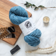 Medium blue woolen-spun fingering yarn arranged with dried plants, a jawbone, a candle, feathers and charred wood.