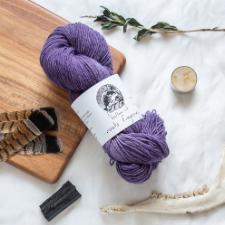 Medium purple woolen-spun fingering yarn arranged with dried plants, a jawbone, a candle, feathers and charred wood.
