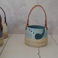 Ceramic yarn bowl with large scoop opening and several small holes. Bowl has a leather strap handle.