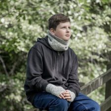 Boy sitting in woods wears cowl and matching mitts.