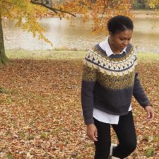 Pullover with geometric colorwork from the neckline halfway to the elbow. Woman wearing the sweater is walking among falling leaves at waterside.