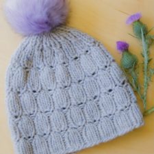 Knitted beanie with seed motif in texture. Thistle-color pom-pom on top. Beanie is shown next to cut thistle flowers.