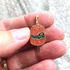 Enamel pin of skein with crochet hook sticking through it, with the word Crochet.