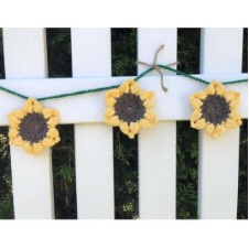 Crocheted sunflowers about the size of a hand strung on a line across a picket fence.