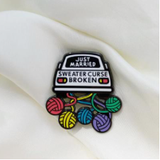 Enamel pin of a car dragging several balls of yarn like tin cans. On the car is written, Just married. Sweater curse broken.