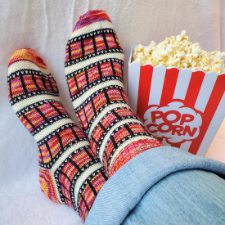 Colorwork socks in alternating stripes, where one stripe looks like a strip of movie film. Next to the feet wearing these cuties is a box of popcorn.