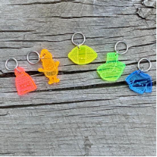 Neon Lucite stitch markers that are characters in the form of a knitted beanie, a skein, a lemon, a pair of socks and a pullover.