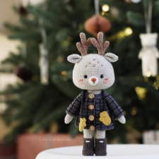 Crocheted reindeer in short coat and boots, with mittens on a string. Ellie is standing in front of a Christmas tree.