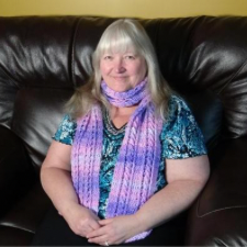 Designer sits on a large leather sofa wearing a blue floral shirt and a solid color crocheted scarf with various cables.