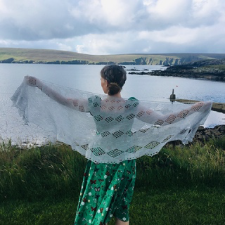 Woman on lake shore facing the lake holds open lace crescent shawl with diamond motifs on a garter stitch background.