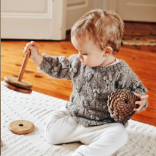 Toddler plays with wooden toy and pine cone, while wearing long sleeve crewneck pullover with bobbles and lace.
