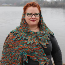 Woman by body of water wrapped in shawl with short row petal design in two colors. Effect is reminiscent of a dragon queen.