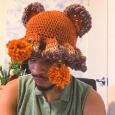 Crocheted hat has fuzzy bear ears and a floppy ruffle border with two pom poms hanging down.