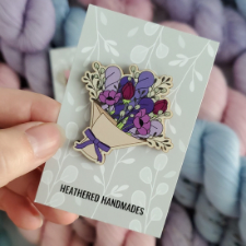 Enamel pin has large, paper-wrapped bouquet of purple flowers and purple skeins.