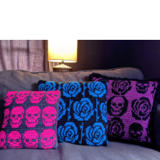 Three pillows, one with pink skulls against black, one with blue roses on black and one with purple and black skulls and roses on purple.