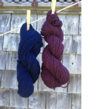 A deep wine skein and a deep blue skein are suspended from a clothesline with wooden clothespins. In the background is a wall covered with weathered wood shakes.