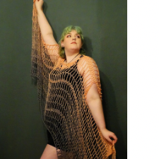 Woman holds aloft the edges of a long netting-style cloak. The cloak has openings for the head and arms to poke through and the “sleeves” continue in an asymmetric shape to the floor.
