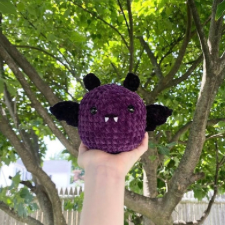 Hand holds aloft a round softball size creature of the night with velour purple body, undersize black wings, black ears, black eyes and tiny white fangs.