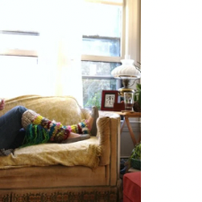At the end of a tan sofa is the lower half of someone wearing jeans, slippers and brightly colored legwarmers made from handspun. In the background is a framed Colonial style papercut and an oil lamp.