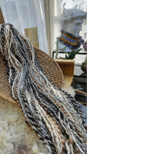 Handspun marled yarn in blacks, grays, whites and brown is draped over a flat basket. In the background is a white bust of a man wearing a quilted cowl.