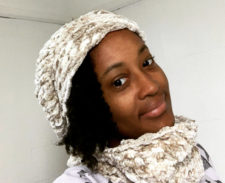 Tania looks over her shoulder smiling slightly, wearing a knitted hat and matching cowl.